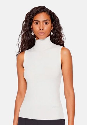 Femme aux longs cheveux bouclés portant un haut sans manches blanc à col roulé et des boucles d'oreilles en forme de goutte, debout devant un fond uni.