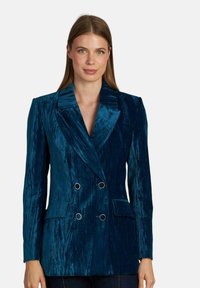 Blazer doppiopetto in velluto teal con revers, due tasche frontali e bottoni circolari in tonalità argento. Tessuto testurizzato con leggero luccichio.