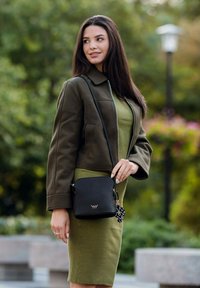 Olivově zelené žebrované šaty spojené s tmavě zelenou bundou. Černá kabelka přes rameno s texturovaným povrchem a detailem loga. Venkovní prostředí.