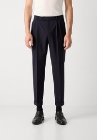 Navy trousers featuring a pleated front, a button closure, and rolled cuffs. Made from a smooth fabric, paired with black loafers and socks.