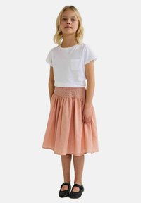 T-shirt blanc à manches courtes avec poche, associé à une jupe de couleur pêche, de longueur genou, avec une taille smockée et un design évasé. Chaussures noires.