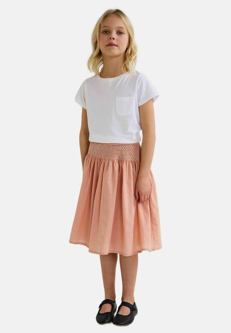 T-shirt blanc à manches courtes avec poche, associé à une jupe de couleur pêche, de longueur genou, avec une taille smockée et un design évasé. Chaussures noires.