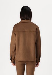 Pullover marrone, oversize, in tessuto morbido con colletto alto, maniche lunghe e una cucitura sulla schiena. Abbinato a pantaloni slim-fit coordinati.