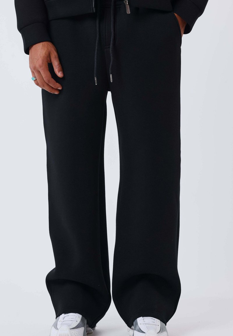 Personne portant un pantalon noir ample avec un cordon de serrage et des baskets blanches, debout avec la main gauche visible et une bague turquoise au doigt.
