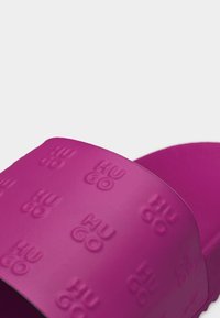 Gros plan sur une bride de sandale à glissière violette vibrante, embossée à plusieurs reprises du nom de la marque "HUGO" en lettres en relief.