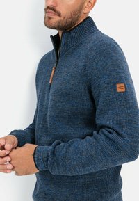 Blue sweater with a high collar and a half-zipper. Knitted texture with a heathered pattern. Leather logo patch on the sleeve.