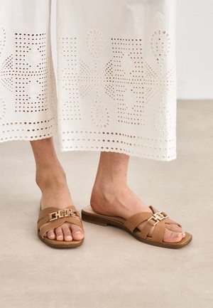 Brown slide sandals with a flat sole, featuring cross straps and a metallic buckle detail. Worn with a white embroidered skirt.