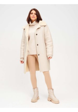 Manteau long beige avec un col en fausse fourrure, détails de boutons, superposé à un léger pull et à des leggings beiges, associé à des bottines crème.