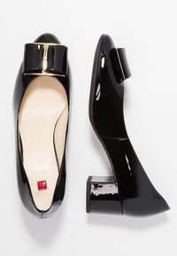 Svarta lackskinnsskor med låg klack, fyrkantig tå, dekorativ rosettdetalj och beige innersula med röd fyrkantig logotyp.
