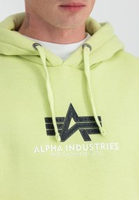 Sudadera con capucha de color verde claro, con un logo negro texturizado y texto blanco, que incluye cordones y ojales metálicos. Textura de tela suave.