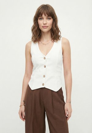 Młoda kobieta o falujących brązowych włosach nosi białą kamizelkę na guziki, brązowe spodnie o szerokich nogawkach oraz złotą biżuterię na jednolitym tle.