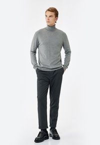 Grauer Rollkragenpullover mit glatter Textur, kombiniert mit dunklen, schmal geschnittenen Hosen und schwarzen Schnürschuhen, vor einem weißen Hintergrund.