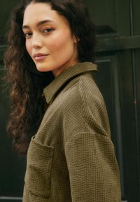 Camicia in velluto a coste verde oliva con colletto, tessuto testurizzato e taschino sul petto, esposta su uno sfondo scuro.