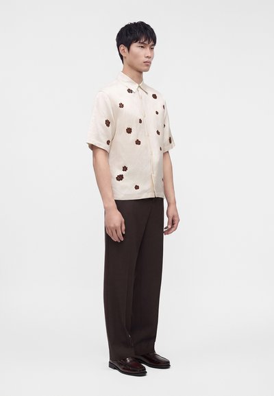 Homme debout portant une chemise beige à manches courtes avec des imprimés floraux marron, un pantalon marron foncé et des chaussures foncées sur un fond uni.