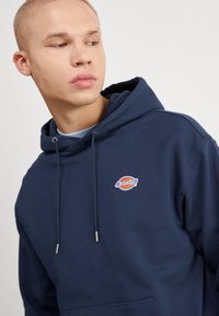 Felpa con cappuccio blu navy, realizzata in tessuto morbido, con tasca frontale e cordini. Include un logo ricamato sul lato sinistro del petto.