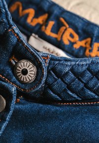 Ceinture en denim avec coutures orange, présentant une texture tressée et un bouton rond en argent orné d'un motif floral. Étiquette visible.