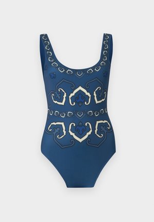 Mladá žena stojaca bosá so skríženými rukami, oblečená v modré vzorovanej jednodielnej plavkách na bielom jednoduchom pozadí.