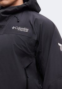 Gros plan d'une personne portant une veste imperméable noire Columbia Titanium avec capuche et poche latérale zippée.