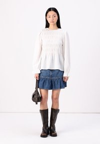Hvit langermet bluse med rynkedetaljer, kombinert med en denim-skirt med lag. Modellen har på seg brune skinnstøvler til kneet og holder en liten veske.