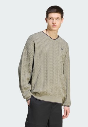 Giovane uomo che indossa un maglione grigio chiaro a trecce con scollo a V e pantaloni neri larghi, in piedi con una mano in tasca, guardando di lato.