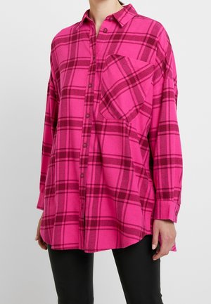 Femme portant une chemise à carreaux ample à manches longues de couleur rose et rouge foncé avec une poche avant, ainsi qu'un pantalon noir, sur un fond uni.