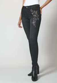 Kvinna klädd i svarta skinny jeans med blommigt broderi på låret och svarta klackiga ankelboots, stående mot en enfärgad bakgrund.