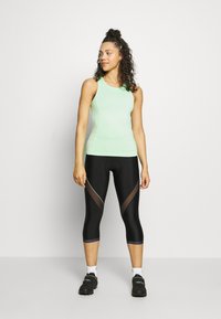 Top sem mangas de cor verde claro com um design ajustado, combinado com leggings capri pretas que apresentam painéis de malha transparente e uma faixa lateral em contraste.