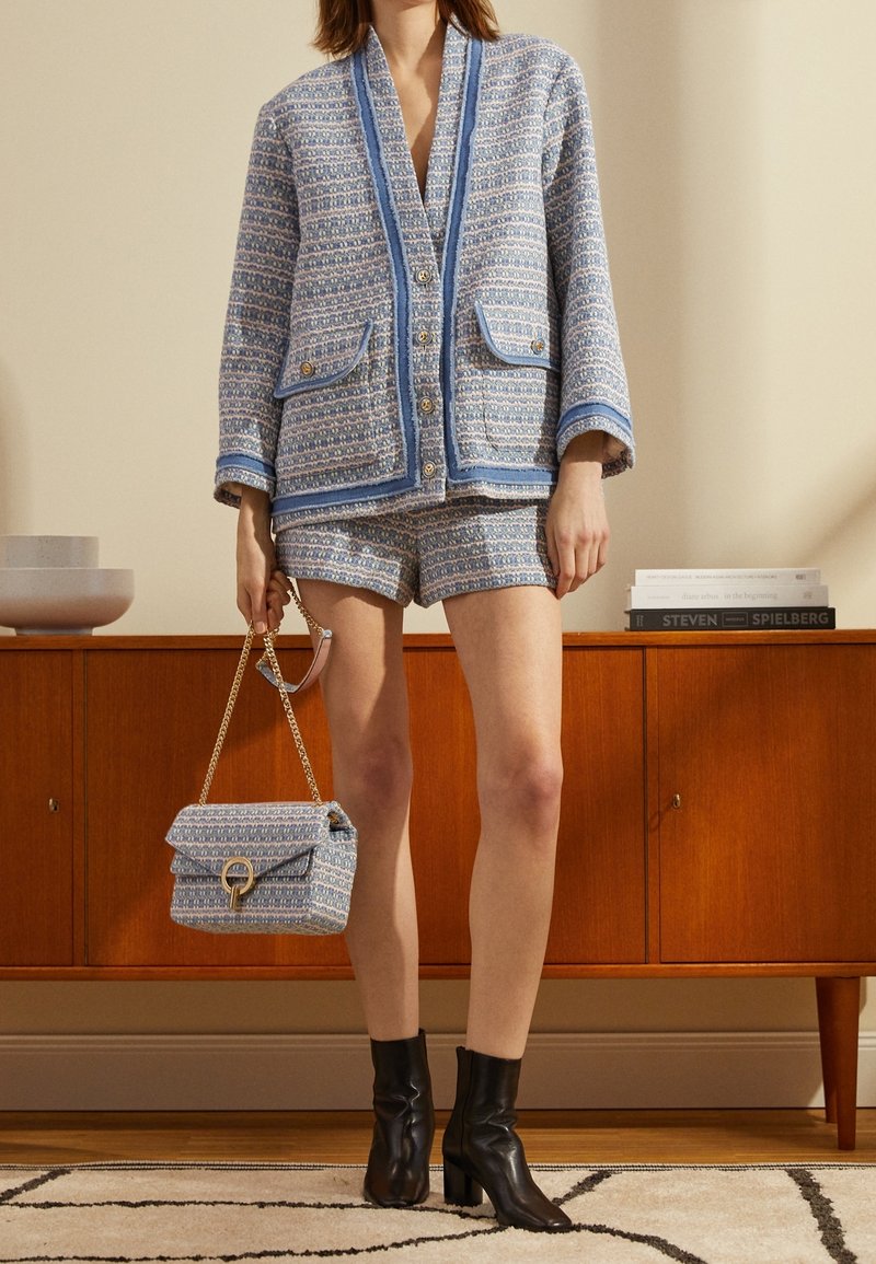 Blå och vit mönstrad vävd jacka med matchande shorts, gyllene knappar och två framfickor; tillsammans med en blå handväska och svarta stövlar.