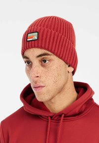 Rode geribde beanie met een genaaid label dat bergen en strepen toont. Gemaakt van zachte gebreide stof. Past snugly op het hoofd.