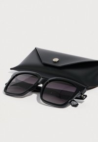 Czarne prostokątne okulary przeciwsłoneczne z ciemnymi soczewkami, wyposażone w grube oprawki i metalowe akcenty, umieszczone obok czarnego skórzanego etui.