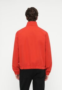 Κόκκινο αδιάβροχο μπουφάν με ψηλό γιακά, ελαστική ύφανση στο τελείωμα και στους καρπούς. Μαλακό ύφασμα, άνετη εφαρμογή, απλός σχεδιασμός και χωρίς ορατές αξεσουάρ.