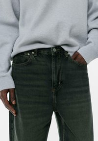 Henkilö, jolla on tummanvihreät farkut ja vaaleanharmaa neule, yksi käsi taskussa ja toinen rentona sivulla.