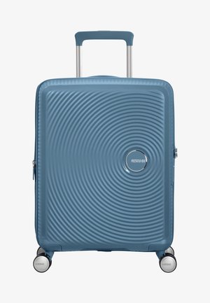 Sininen kovakuorinen matkalaukku, jossa on konsentrisen ympyrän kuviointi, neljä pyörivää pyörää ja jatkettu teleskooppikahva.