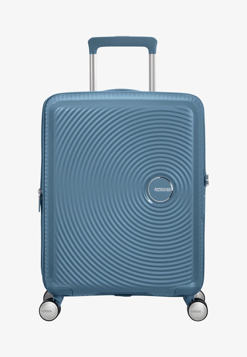 Valise rigide bleue avec texture en cercles concentriques, quatre roues pivotantes et poignée télescopique déployée.