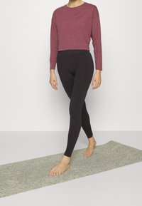 Top cropped a maniche lunghe color ruggine con leggings neri, entrambi realizzati in materiale morbido ed elasticizzato. A piedi nudi, cammina su un tappetino grigio con superficie testurizzata.