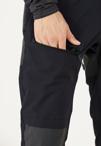 Mustat tekniset housut, joissa on vetoketjullinen tasku reidessä, ominaisuutena sileä pinta ja erottuva harmaa paneeli alhaalla.