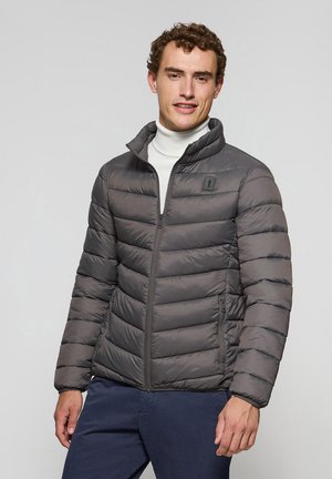 Chaqueta acolchada gris con diseño de acolchado horizontal, cremallera frontal y cuello alzado. El material parece suave y ligero. Dos bolsillos laterales.