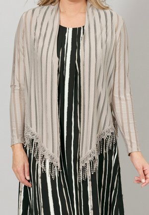 Femme portant un châle à franges beige rayé sur une robe à rayures verticales noire et blanche, debout devant un fond neutre.