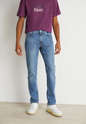Mann trägt blaue Straight-Leg-Jeans, weiße Sneakers und ein bordeauxfarbenes Calvin Klein T-Shirt, steht vor einem einfarbigen hellen Hintergrund.