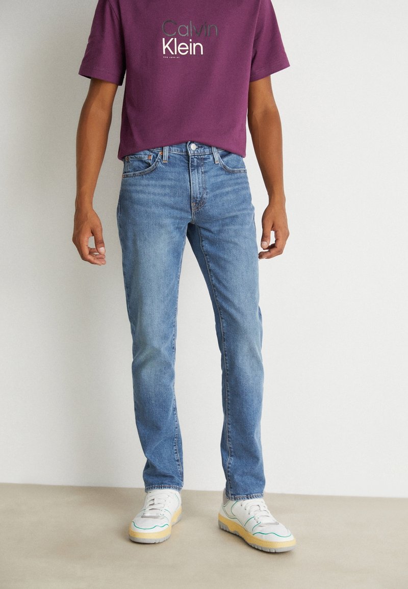 Homme portant un jean droit bleu, des baskets blanches et un t-shirt Calvin Klein bordeaux, debout devant un fond clair uni.