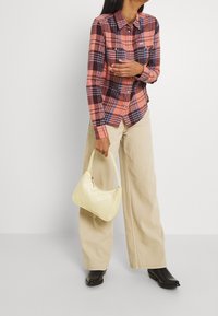 Chemise en flanelle rose à motif à carreaux, pantalon beige à jambes larges, tenant un petit sac à main jaune, assorti à des bottes noires à bout pointu.