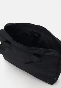 Zwarte stoffen tas met een ritsluiting, twee handvatten en een binnenzak, met een label waarop "TOMMY HILFIGER" staat. Glad textuur.
