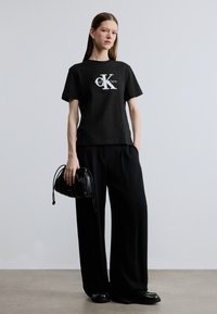 Svart bomulls-T-skjorte med hvit Calvin Klein-logo, kombinert med vide, svarte bukser. Modellen holder en liten svart snorpose.