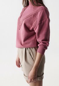 Sudadera rosa con detalles de encaje en un patrón de chevrón, con un cuello alto y mangas largas, combinada con pantalones cortos beige.