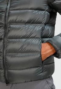 Opgeblazen jacket in donkergrijs nylon. Voorzien van een rits aan de voorkant, horizontale naden en zijzakken met een gladde afwerking.
