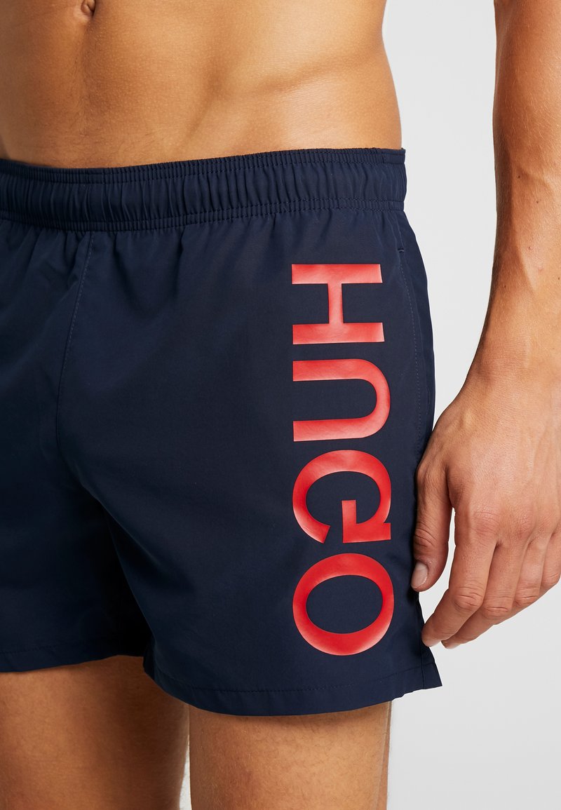 Marineblaue Schwimmshorts mit einem roten Gummilogo "HUGO" auf der linken Seite, ausgestattet mit einem elastischen Bund und einem glatten, leichten Stoff.