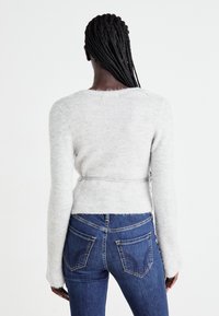 Hellgrauer Pullover mit langen Ärmeln, tailliertem Design und Rundhalsausschnitt, kombiniert mit dunkelblauen Jeans, die detaillierte Gesäßtaschen aufweisen.