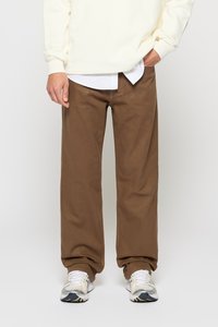Pantaloni in cotone marroni con una vestibilità sciolta, abbinati a una felpa crema e scarpe da corsa grigie. Il tessuto ha una consistenza liscia.