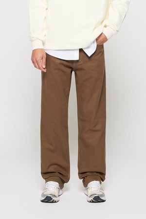 Brune bomullsbukser med løs passform, kombinert med en kremfarget sweatshirt og grå joggesko. Stoffet har en glatt tekstur.