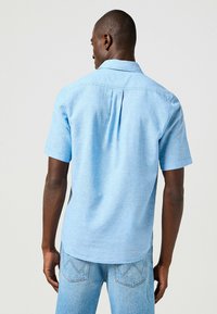 Camicetta a maniche corte, di un azzurro chiaro, in tessuto morbido, con una piega sul retro e colletto a punta, abbinata a jeans in denim azzurri chiari.